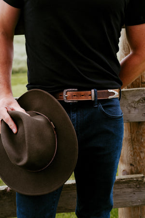Person holding a brown cowboy hat with a wooden fence and grass in the background modeling Dakota Belt in Brown