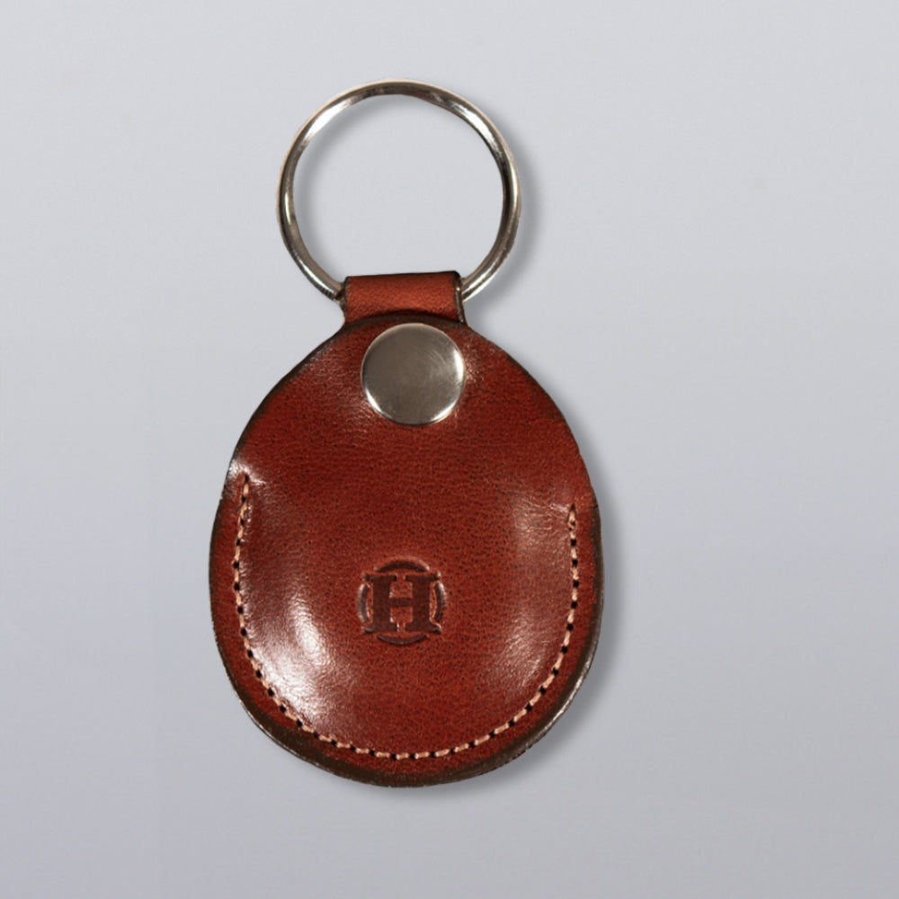 The Circular Tag Holder by Hanks Belts, made of brown full-grain leather with a silver metal keyring and button, features a circular embossed &quot;H&quot; logo and is shown on a light gray background.