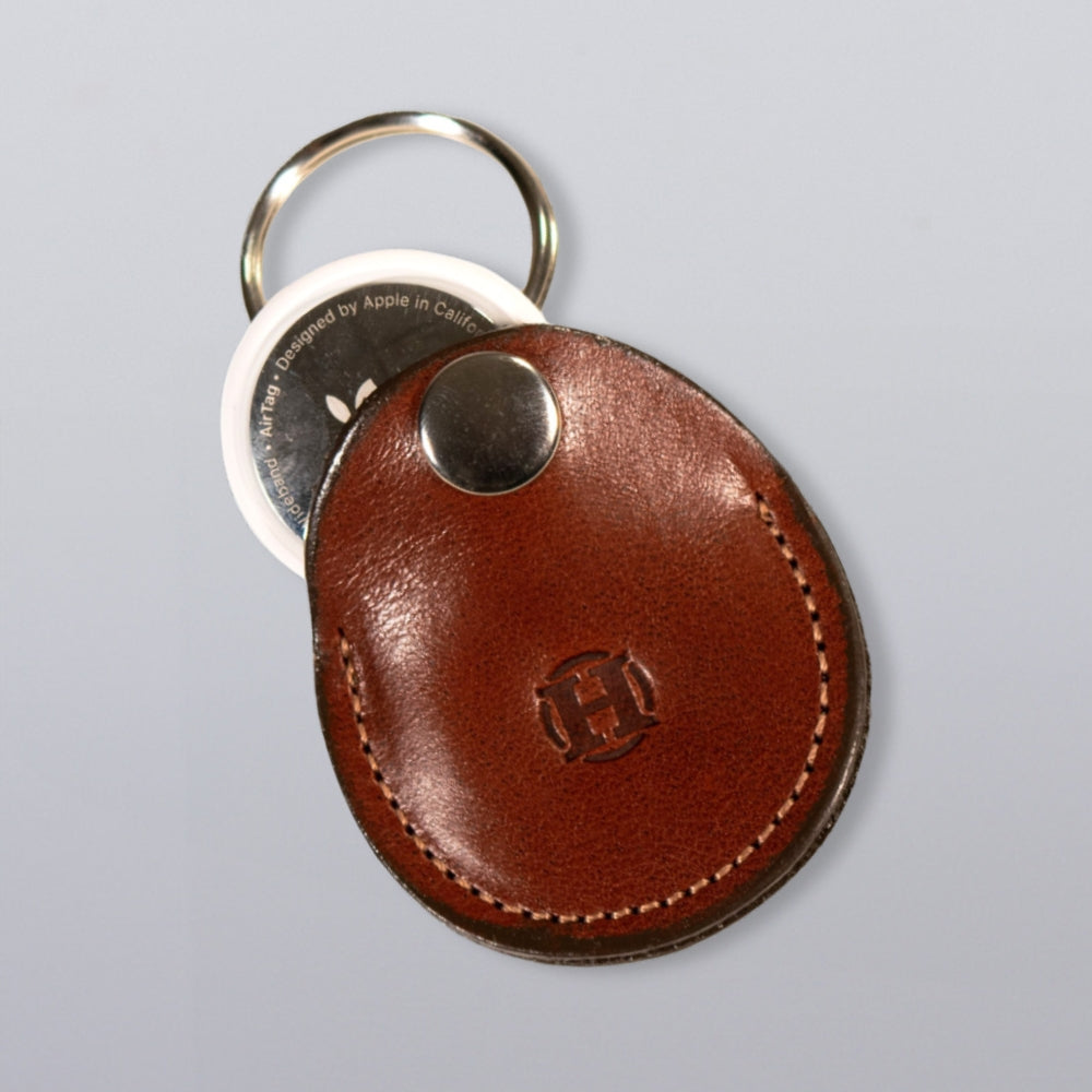 The Hanks Belts Circular Tag Holder, made of brown full-grain leather with stitched borders and an embossed logo, holds a round white Apple AirTag. It attaches to a metal key ring and is shown on a light gray background.