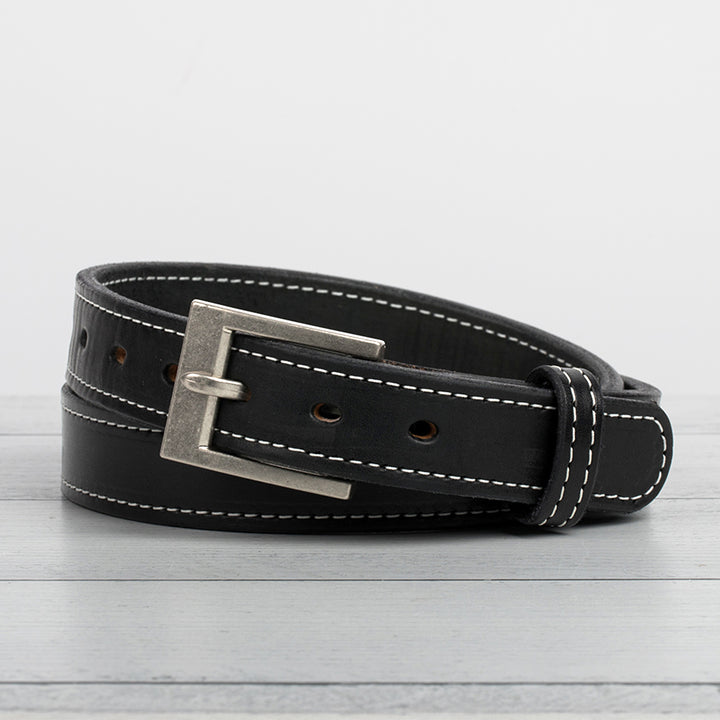 The Bonnie by Hanks Belts is a black full-grain vegetable tanned leather belt with white stitching and a silver rectangular buckle, shown coiled on a light gray wooden surface against a plain white background.