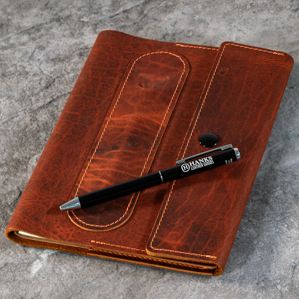 A Luxury Bison Journal from Hanks Belts, crafted in brown leather with visible stitching, lies closed on a gray stone surface. A black pen marked &quot;Hanks Belts&quot; rests diagonally across the journal.