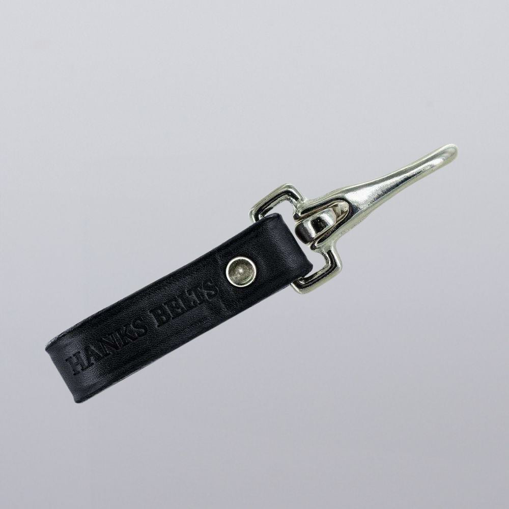 A black leather Canyon Keychain from Hanks Belts features a rugged silver clip and snap button, with &quot;HANKS BELTS&quot; embossed on the strap. The keychain is shown against a plain light gray background.