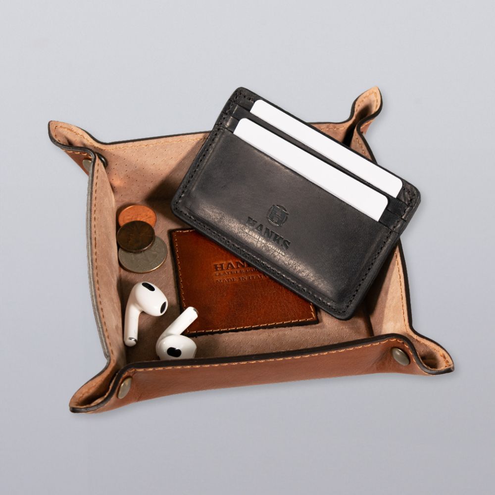 The Italian Compact Valet Tray by Hanks Belts neatly holds a black cardholder with two cards, a brown leather accessory, coins, and wireless earbuds on a light gray surface.