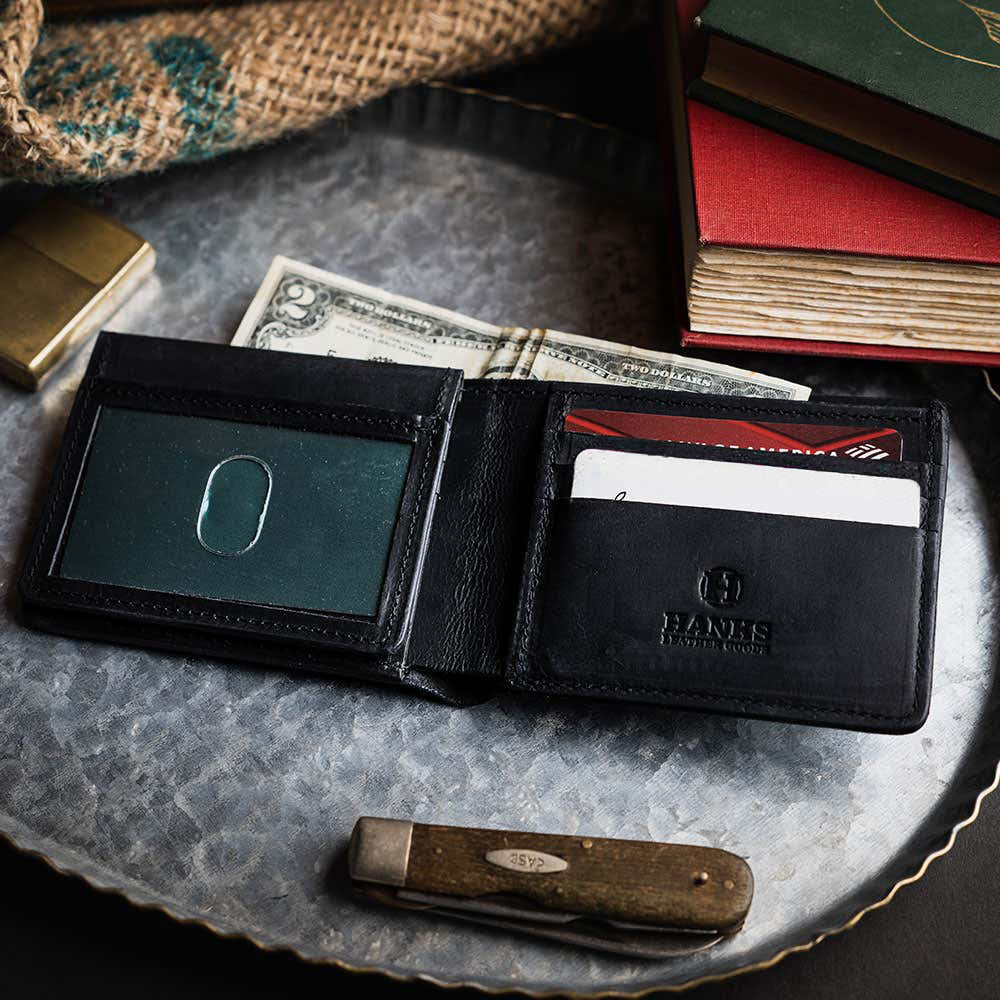 The Huron Premium Bifold with ID Window by Hanks Belts, made in the USA, sits open on a metal tray displaying cards and cash, surrounded by a pocketknife, lighter, books, and a burlap sack.