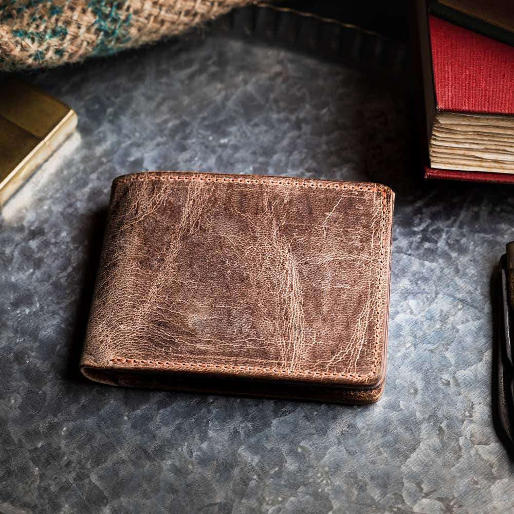 The Hanks Belts Huron Premium Bifold with ID Window, made from brown steerhide leather, sits on a textured metal surface beside a burlap cloth, a closed red book, and part of another dark item.