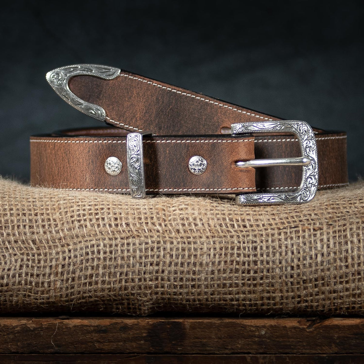 The Willow Belt - 1.25&quot; by Hanks Belts, coiled neatly on a rustic burlap cloth, showcases intricate silver buckle and tip details. The floral buckle and two decorative silver buttons feature Western-style engravings. The dark background highlights the craftsmanship of this brown leather belt, which comes with a 100-year warranty.