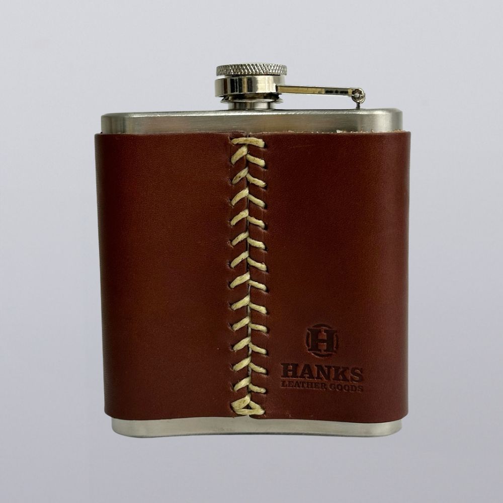 The Leather Covered Flask by Hanks Belts, made in the USA, features brown leather with white stitching and an embossed logo on front. This stainless steel flask is displayed against a plain light background.