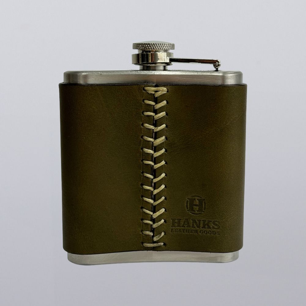 Made in the USA, the Hanks Belts Leather Covered Flask is a stainless steel flask wrapped in olive green leather with beige stitching, a screw cap, and the &quot;HANKS BELTS&quot; logo stamped on the front.