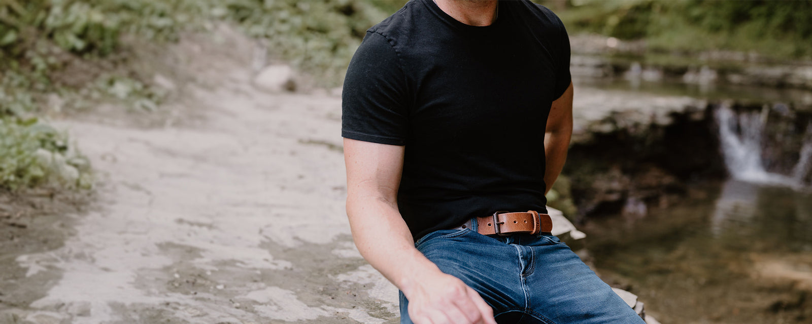 Male model sitting on a rock outdoors wearing Hanks Rustic Glaze Belt with jeans and a black tee
