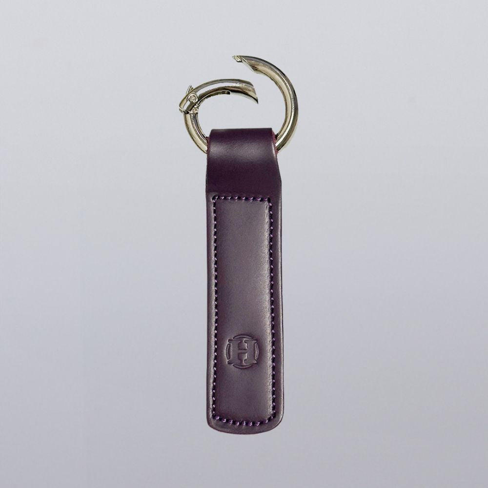 The Superior Keychain by Hanks Belts is a purple leather rectangle with visible stitching and an embossed logo near the bottom, featuring a silver open gate keyring at the top, set against a plain light gray background.