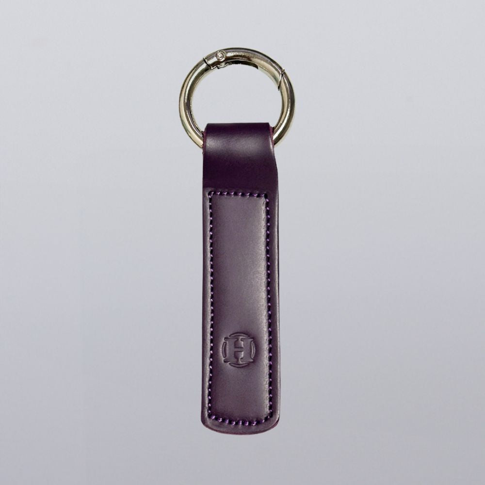 The Hanks Belts Superior Keychain is a purple leather rectangle with visible stitching and an embossed monogram near the bottom. It features a sleek silver open gate keyring set against a plain light gray background.