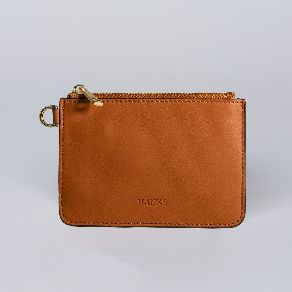 The Livia Mini Wristlet by Hanks Belts is a small, rectangular brown leather pouch with a gold zipper, metal side ring, and "HANKS" embossed on the front, displayed against a plain light gray background.