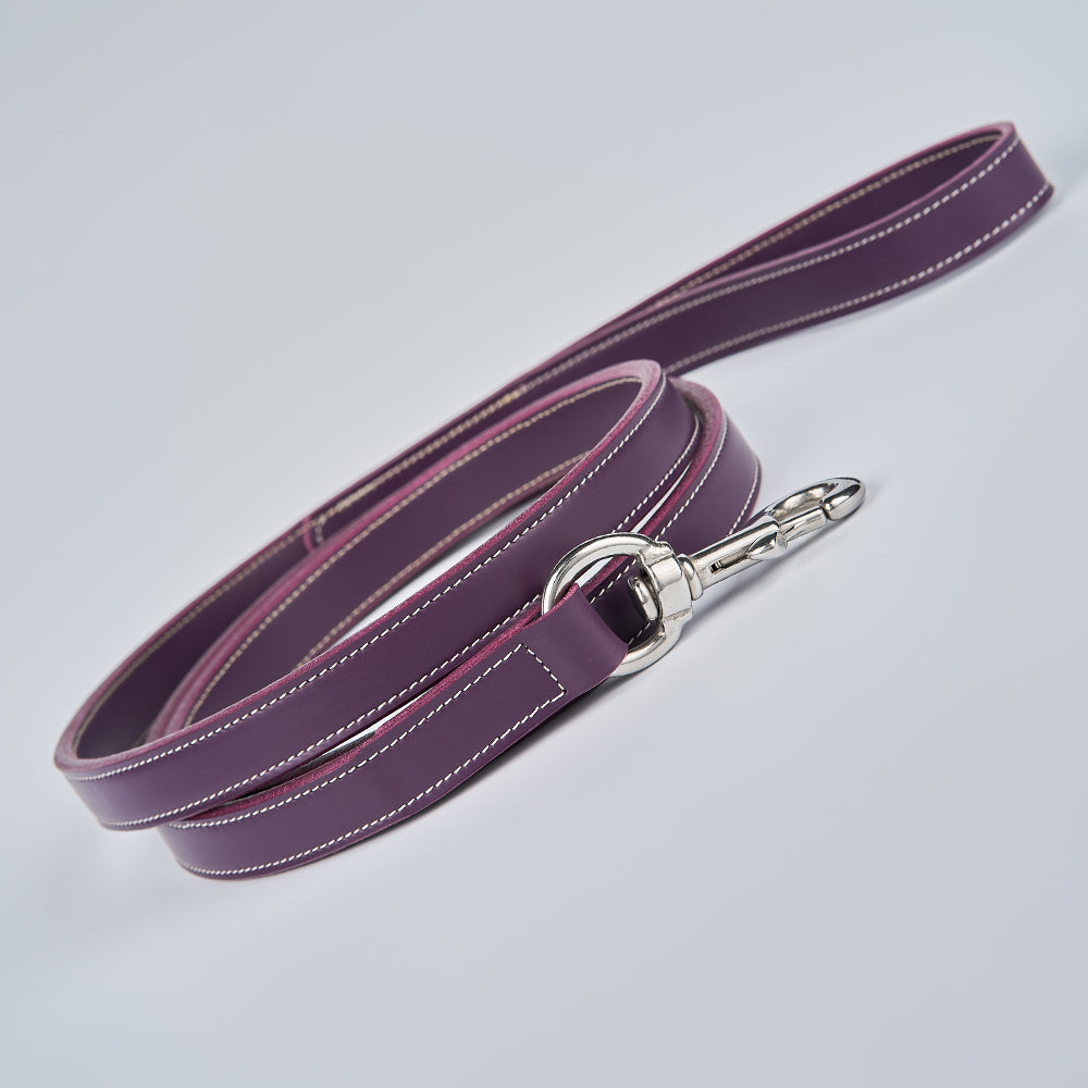 The Hanks Belts Old Reliable Leash is a coiled purple English bridle leather dog leash with white stitching and a silver metal clasp, displayed on a light gray surface. Made in the USA.