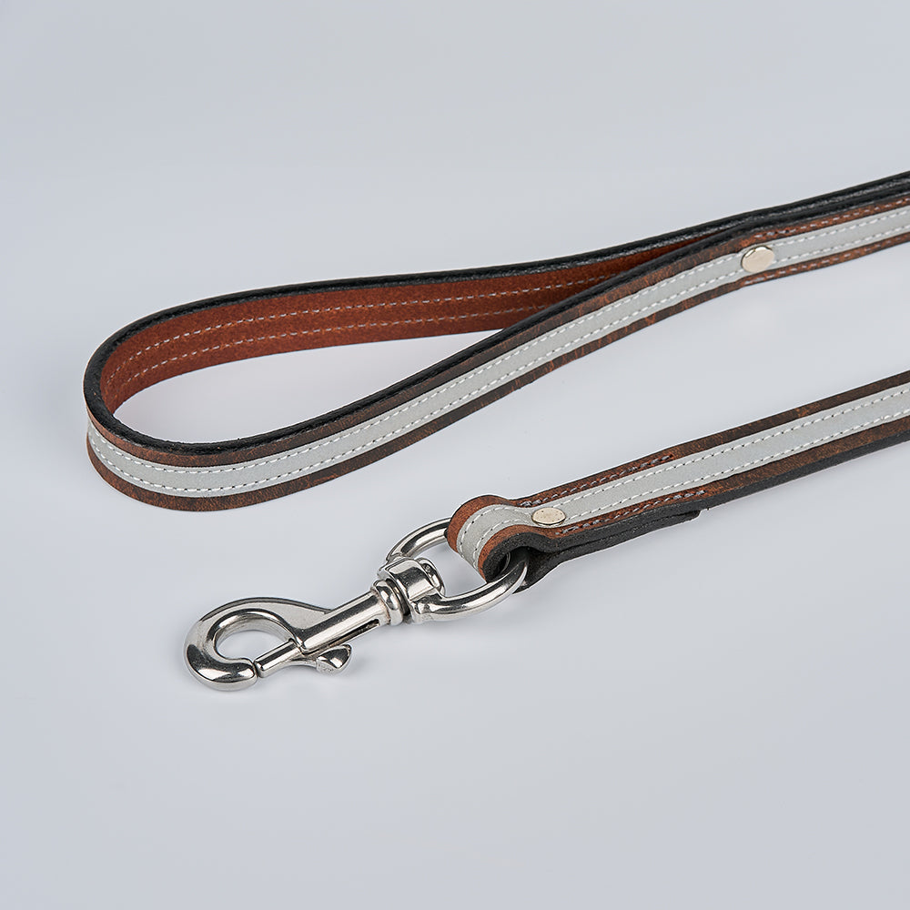 The Remi Reflective Leash by Hanks Belts features brown bison leather, white stitching, and a silver clip—coiled on a light gray background—offering a secure grip and visibility for safe night dog walks.
