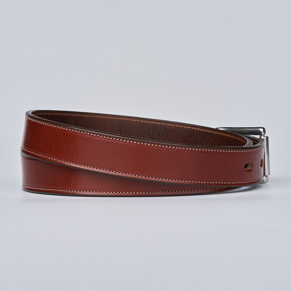 The Highland Lined by Hanks Belts, crafted from brown English bridle leather with white stitching, is elegantly coiled on a light gray background. Its silver buckle peeks out, emphasizing the premium quality of this refined accessory.