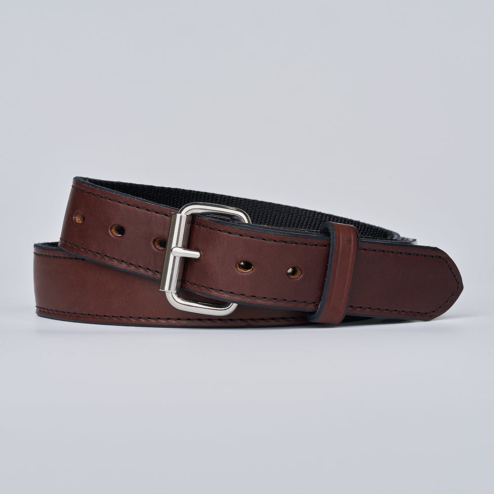 The Kydex Reinforced by Hanks Belts, a brown leather belt with a silver rectangular buckle, is coiled on a light gray background. It features visible edge stitching, multiple adjustment holes, and a rugged design for daily use.