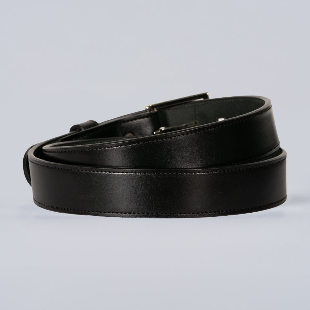 The Ledger by Hanks Belts is a black leather belt with a simple, polished finish and a silver buckle, displayed coiled neatly on a light gray surface.