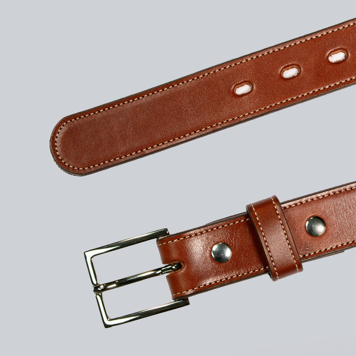 The Ledger by Hanks Belts, a brown leather belt with a silver buckle, is displayed on a light gray background. The stitching and adjustment holes are clearly visible.