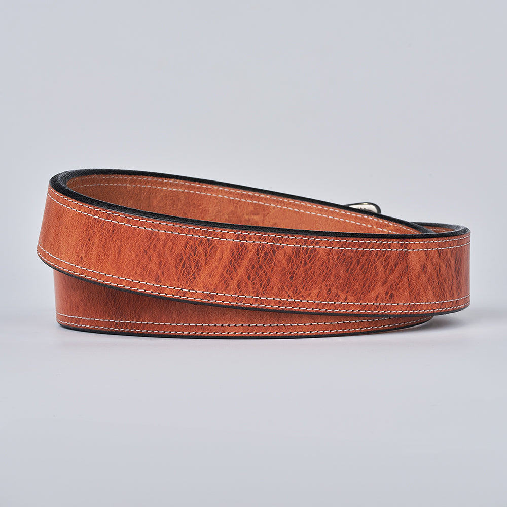 The Old World Harness by Hanks Belts, crafted from USA-tanned Hermann Oak full-grain leather, features visible texture and white edge stitching. Shown coiled on a light gray background.