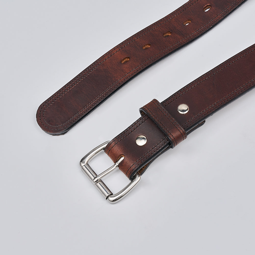 A close-up of The Old World Harness by Hanks Belts, made from USA-tanned Hermann Oak full-grain leather with a silver buckle and metal rivets, lies flat on a light gray surface, displaying multiple adjustment holes.