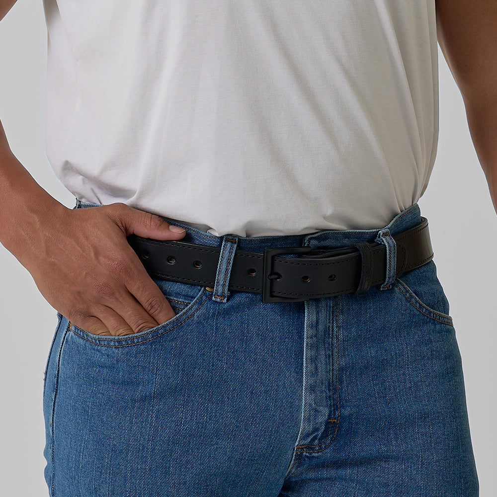 A person wears a white t-shirt tucked into blue jeans with The Black Out Tactical CCW belt by Hanks Belts, one hand in their front pocket, against a plain light background.