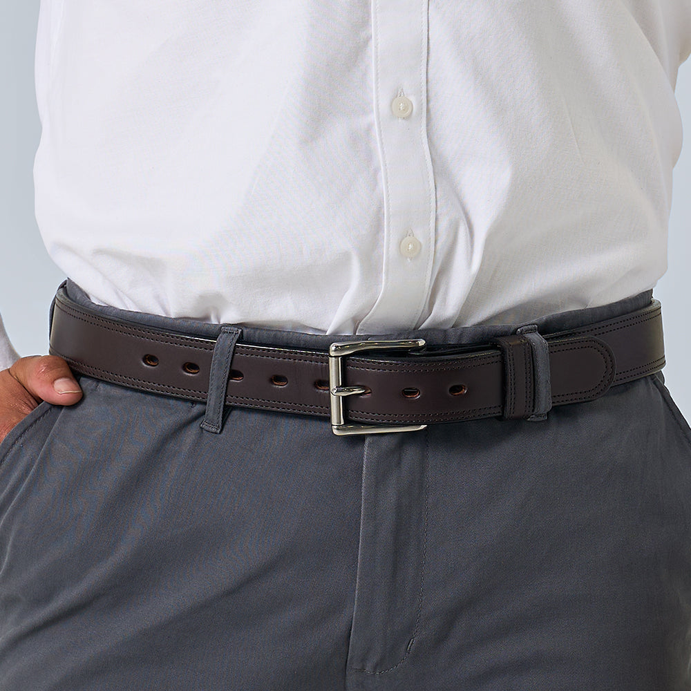 A person wears a white button-down shirt with gray pants, secured by Hanks Belts&#39; The Brute Double Layer dark brown vegetable tanned leather belt with a silver buckle. One hand is in their pocket.