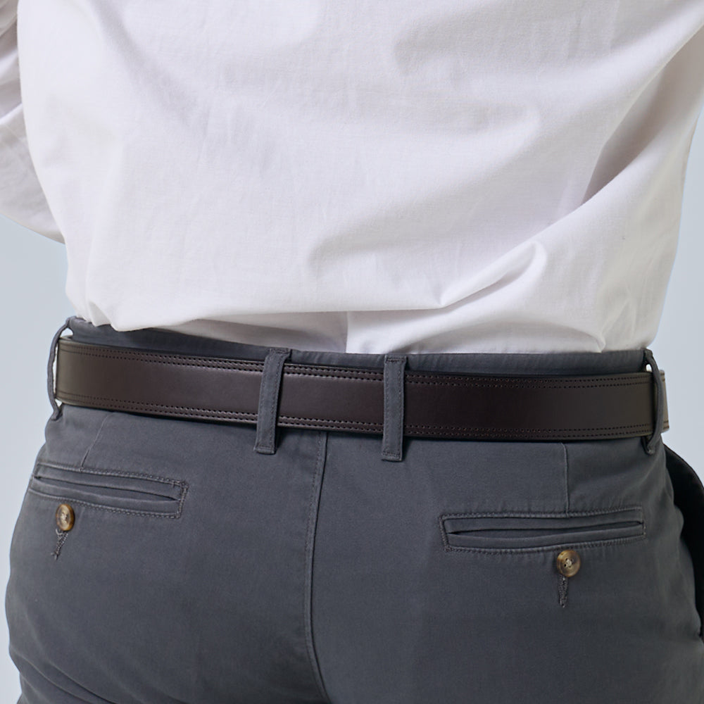 A person shown from the back wears a white shirt, gray pants with buttoned back pockets, and The Brute Double Layer belt by Hanks Belts in dark brown vegetable tanned leather.