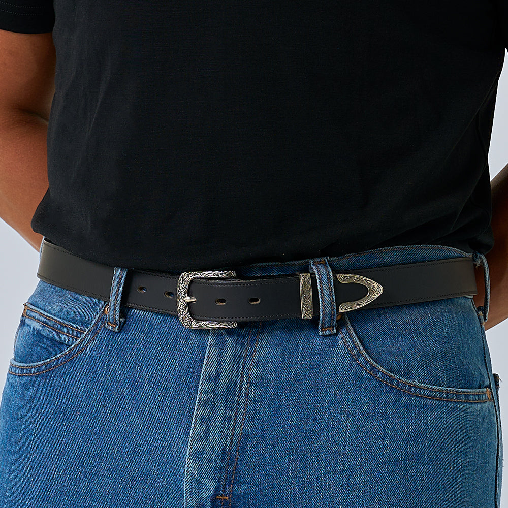 A person wearing a black t-shirt tucked into blue jeans with Hanks Belts&#39; The Dakota Bison western belt, crafted from bison leather and featuring a decorative floral buckle set with matching silver belt tip.