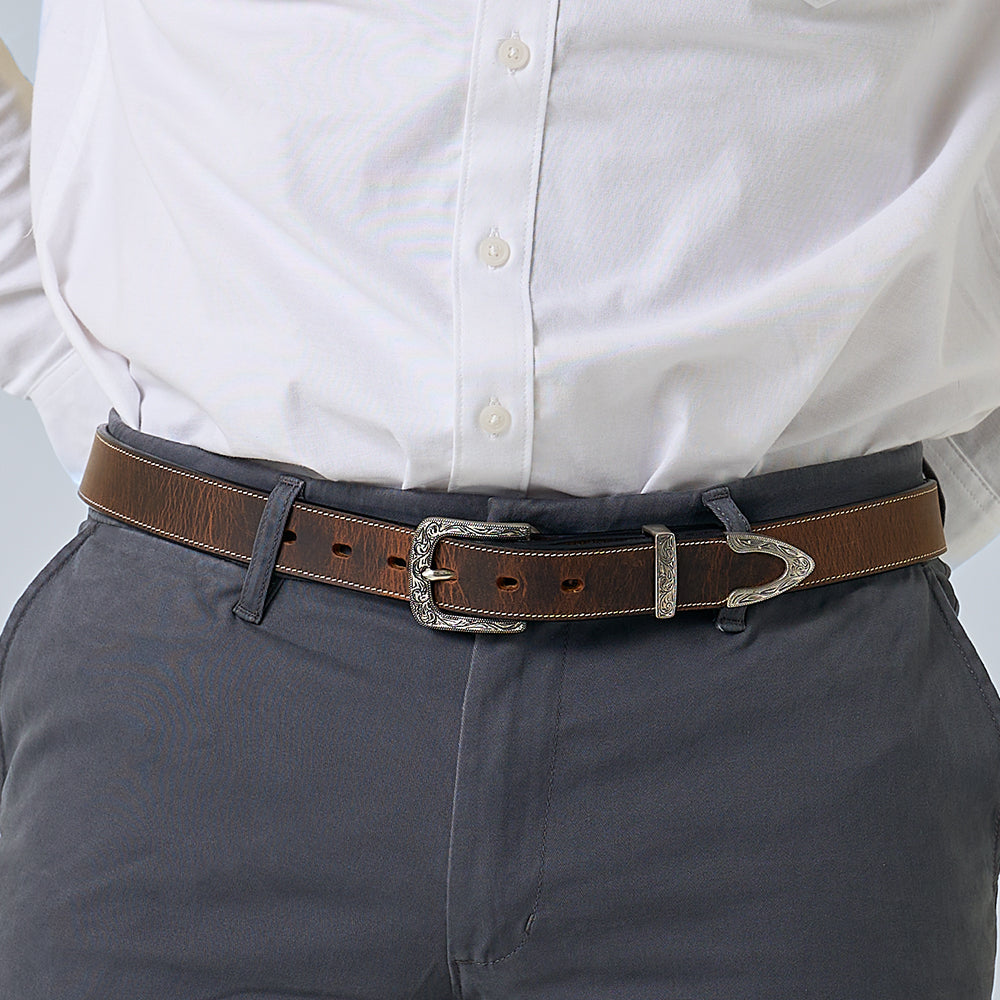 A person wearing a white button-up shirt tucked into dark gray pants with The Dakota Bison belt by Hanks Belts, featuring a decorative silver floral buckle set and tip.