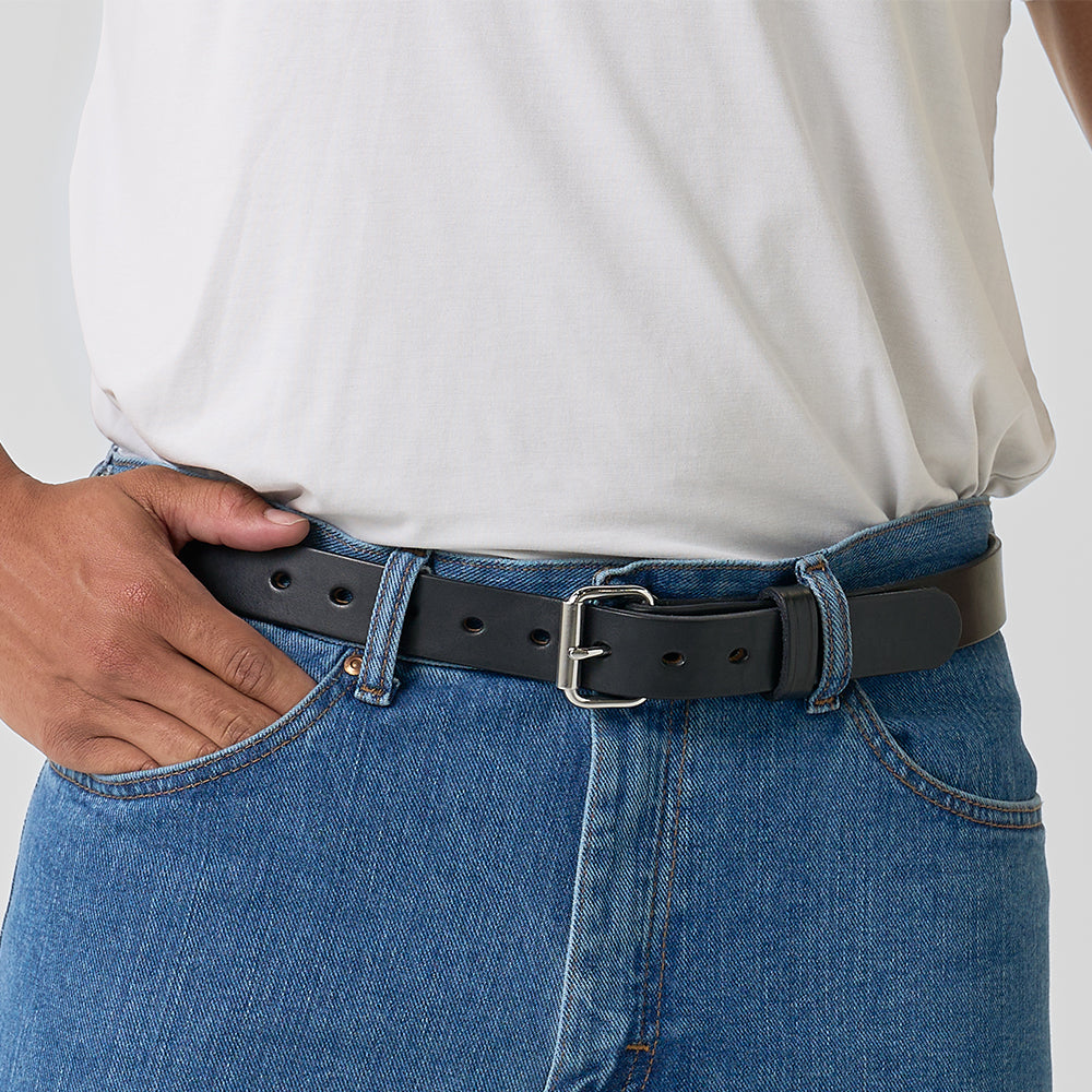 A person in a white t-shirt and blue jeans wears Hanks Belts&#39; The Deputy, a black leather concealed carry belt with a silver buckle, threaded through the jean loops and held at the waistband with one hand.