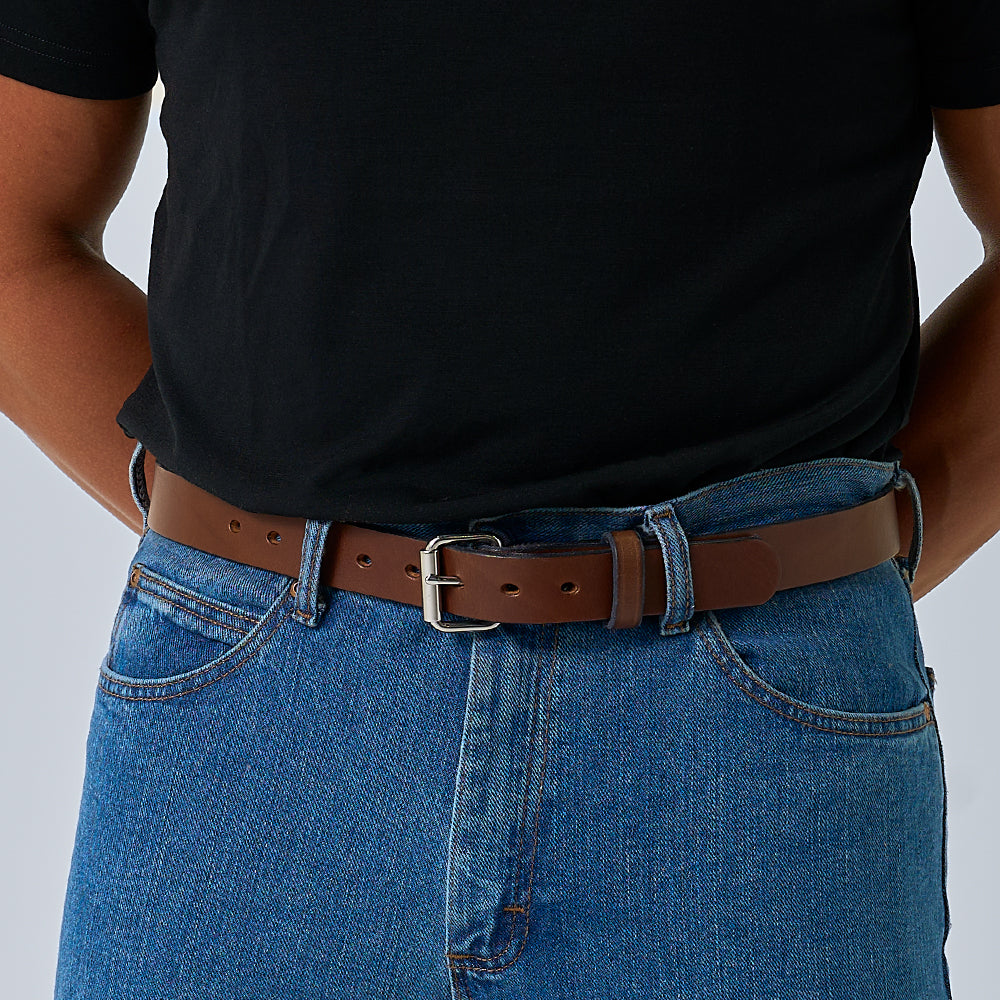 A person stands with hands behind their back, wearing a black t-shirt tucked into blue jeans and &quot;The Deputy&quot; brown leather gun belt by Hanks Belts.