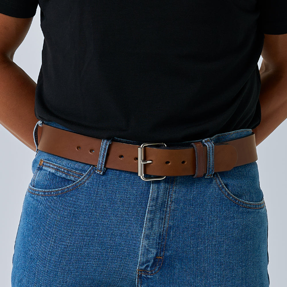 A person in a black t-shirt and blue jeans wears Hanks Belts&#39; &quot;The Enforcer,&quot; a brown 1.75&quot; CCW leather gun belt with a silver buckle. The image is cropped to display only the torso and upper legs, hands positioned behind their back.