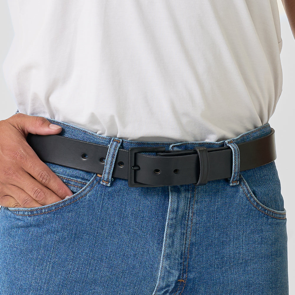 Close-up of a person in blue jeans and a white tucked-in t-shirt, wearing Hanks Belts’ The Everyday— a durable men&#39;s full-grain leather belt with a rectangular buckle. Their left hand rests in their pocket.