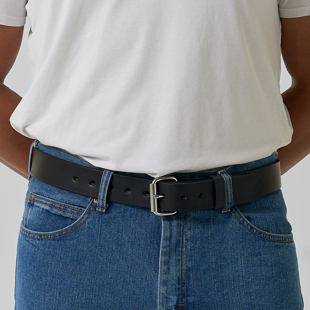 A person wears a plain white T-shirt tucked into blue jeans, secured with The Gunner by Hanks Belts, featuring a silver buckle. Their hands are behind their back.