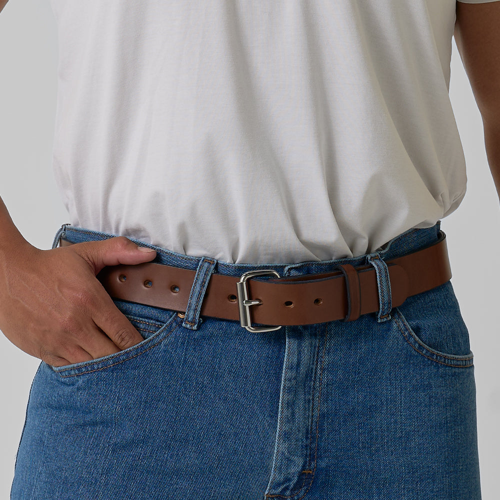 A person wearing a white t-shirt, blue jeans, and The Gunner belt by Hanks Belts with a silver buckle, one hand resting in a pocket.