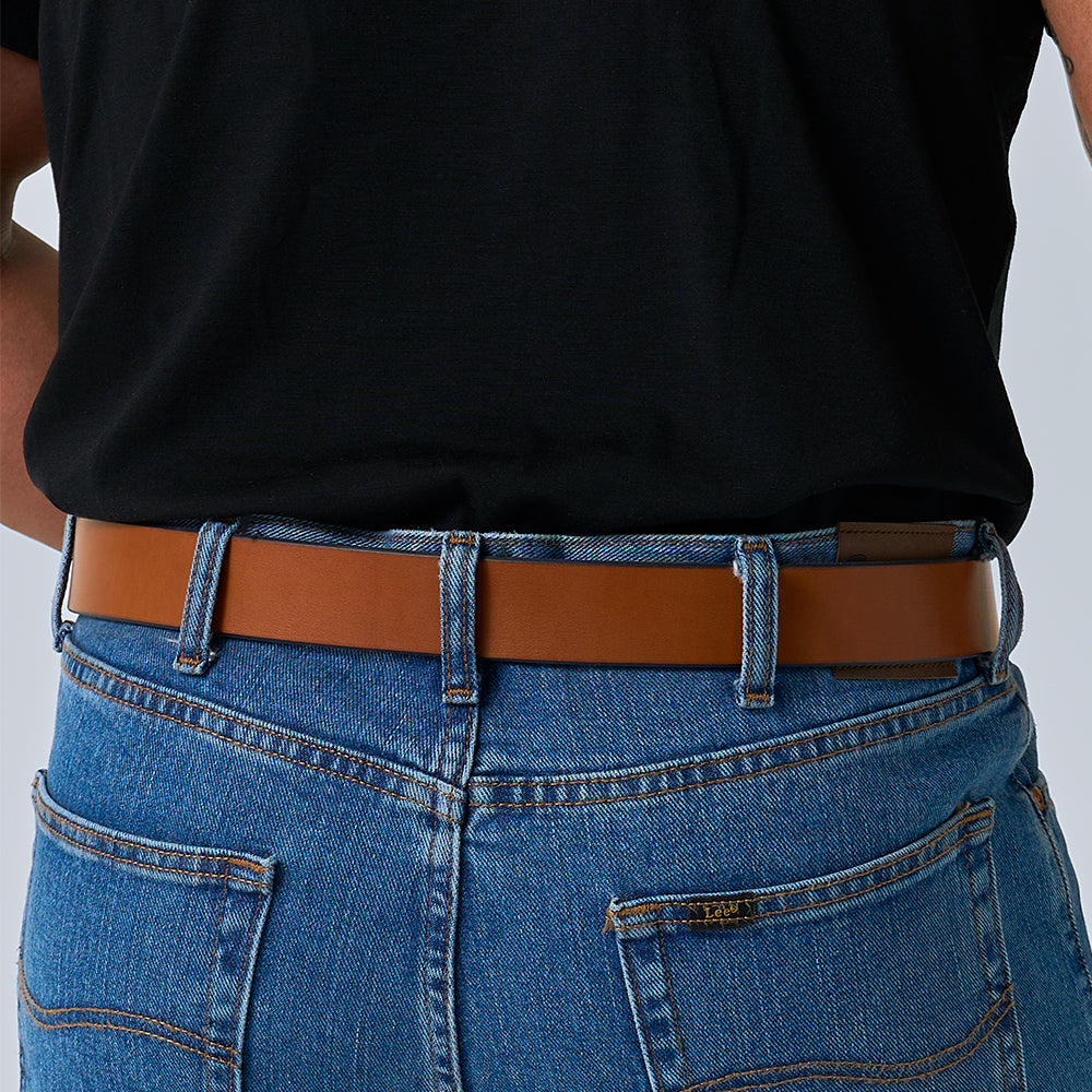 A person wearing blue jeans with visible back pockets and a light brown Hanks Belts &quot;The Gunner&quot; belt, paired with a tucked-in black shirt. The image is cropped to show only the waist and torso.