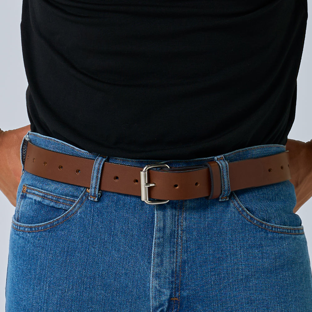 A person wears a black top with blue denim jeans, accessorized with Hanks Belts&#39; The Gunner Utility brown leather belt featuring a silver buckle, hands resting on hips.