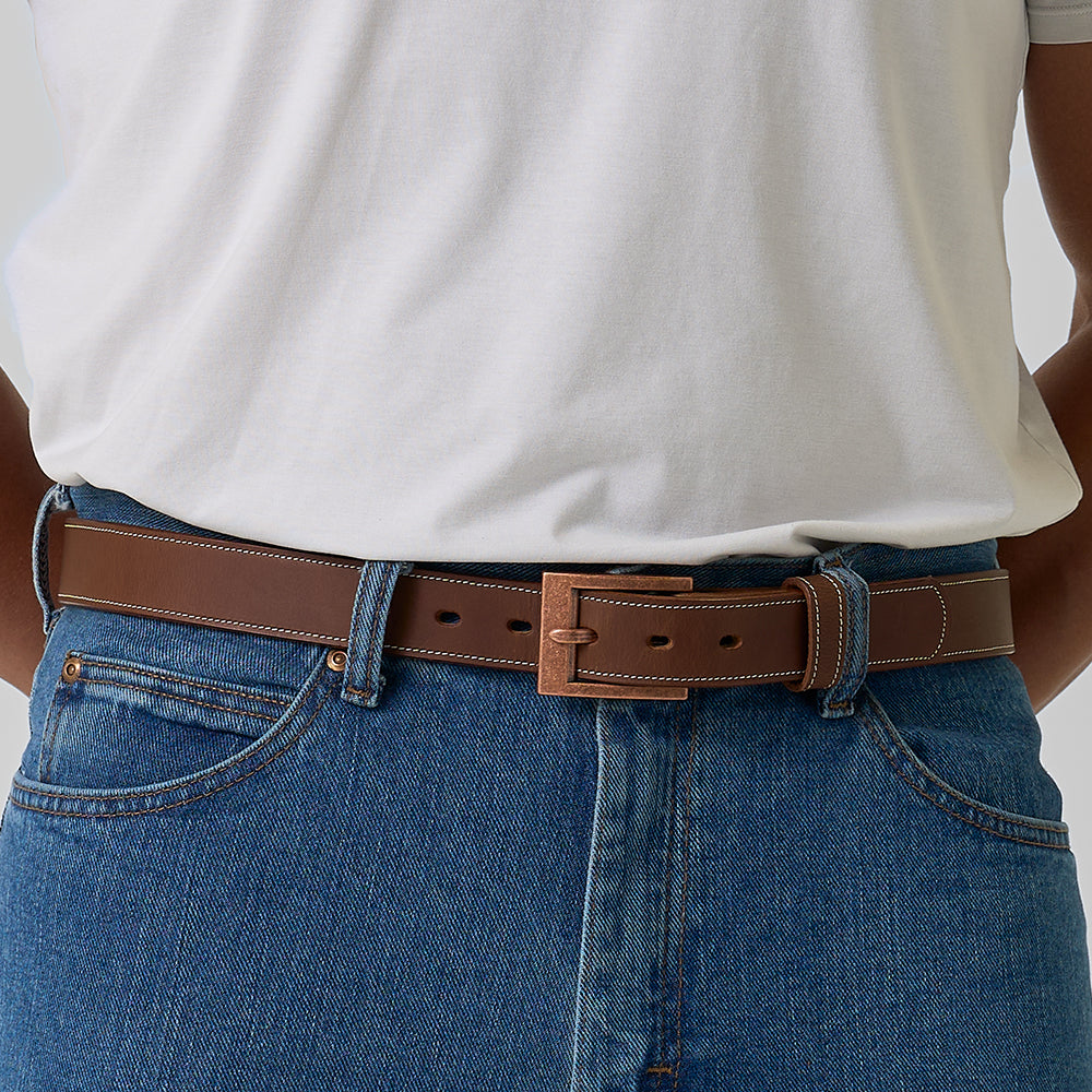 The Heritage Bison by Hanks Belts: a rugged brown bison leather belt with white stitching and a rectangular copper-colored buckle, coiled neatly on a light gray background—showcasing timeless American craftsmanship.