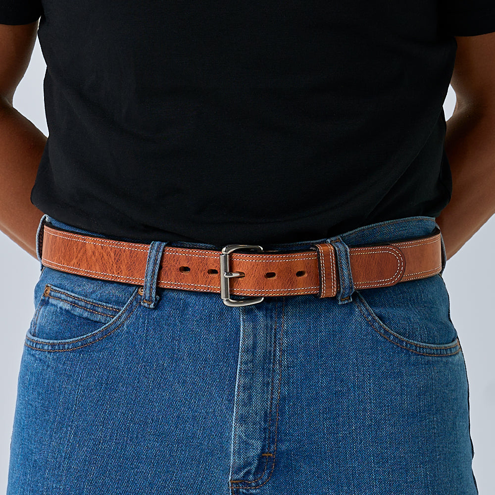 A person wears a black t-shirt tucked into blue jeans, secured with Hanks Belts&#39; The Old World Harness—a brown full-grain leather belt with a silver buckle. Their hands are positioned behind their back.