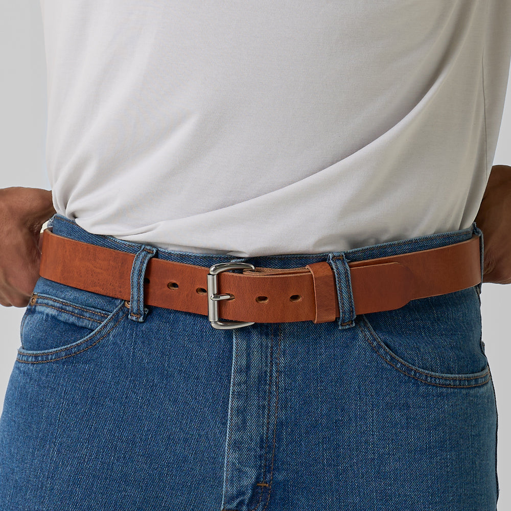 A person shown from the waist down, wearing blue jeans and a white t-shirt, with The Old World Harness full-grain leather belt by Hanks Belts fastened through the belt loops.