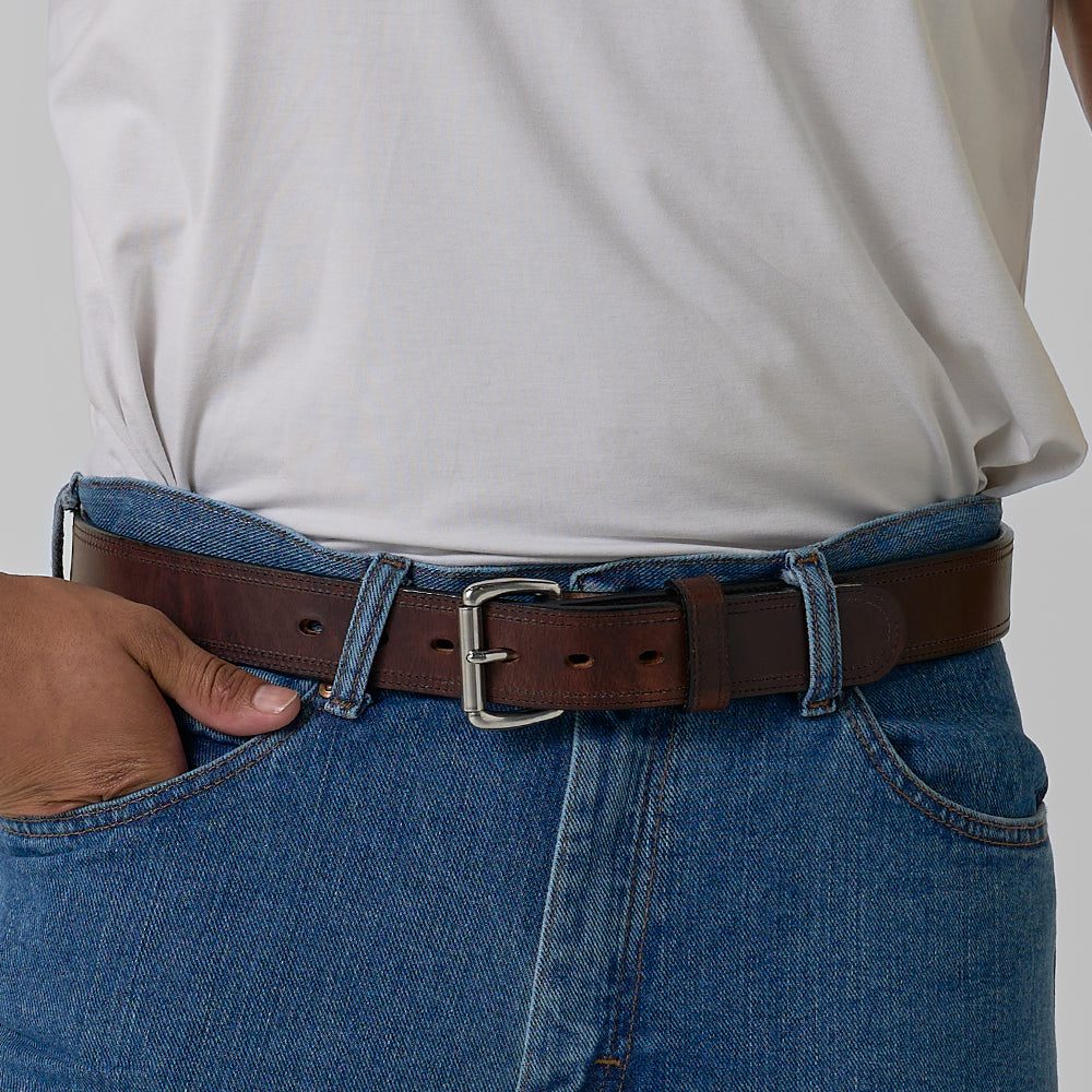 A person wears a white shirt tucked into blue jeans with Hanks Belts&#39; The Old World Harness full-grain leather belt featuring a silver buckle, and one hand rests in a front pocket of the jeans.