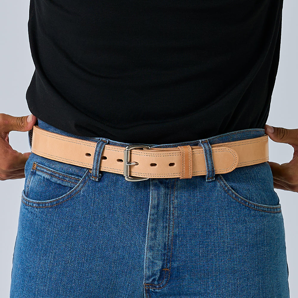 A person in blue jeans and a black shirt wears the Hanks Belts Premier Double Layer, a tan English bridle belt with a silver buckle, with their hands resting on either side of the belt.