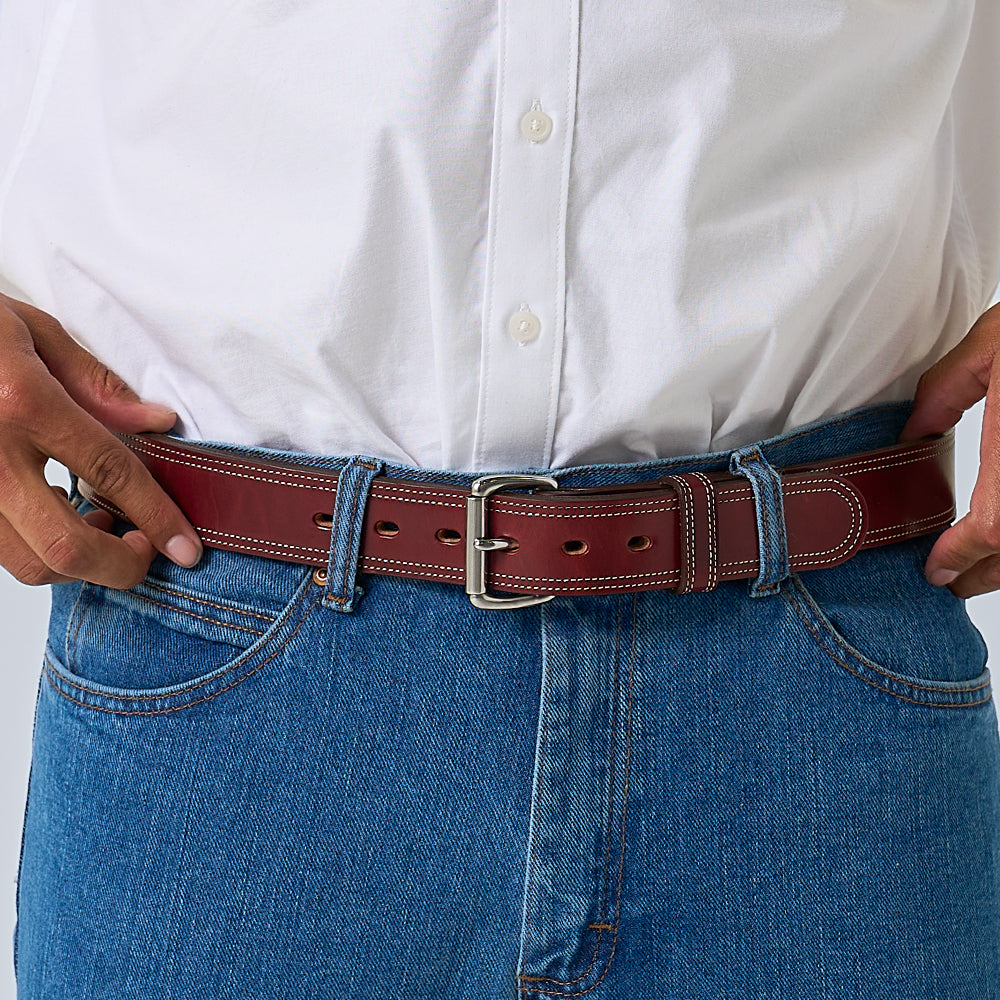 A person in a white button-up shirt and blue jeans fastens The Premier Double Layer by Hanks Belts—a vegetable tanned leather belt with white stitching and a silver buckle.