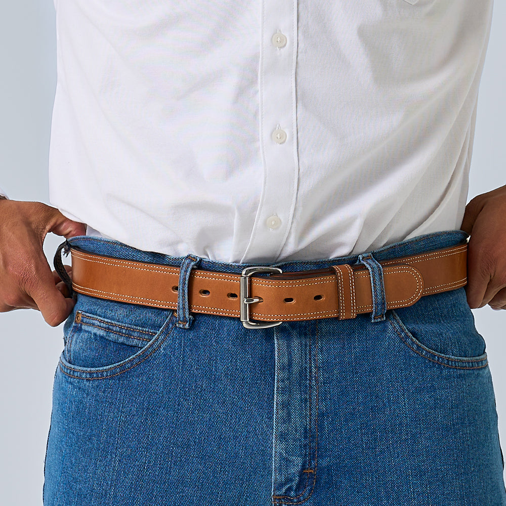 A torso and hands are shown adjusting the waistband of blue jeans with a Hanks Belts The Premier Double Layer vegetable-tanned leather belt, paired with a tucked-in white button-up shirt.
