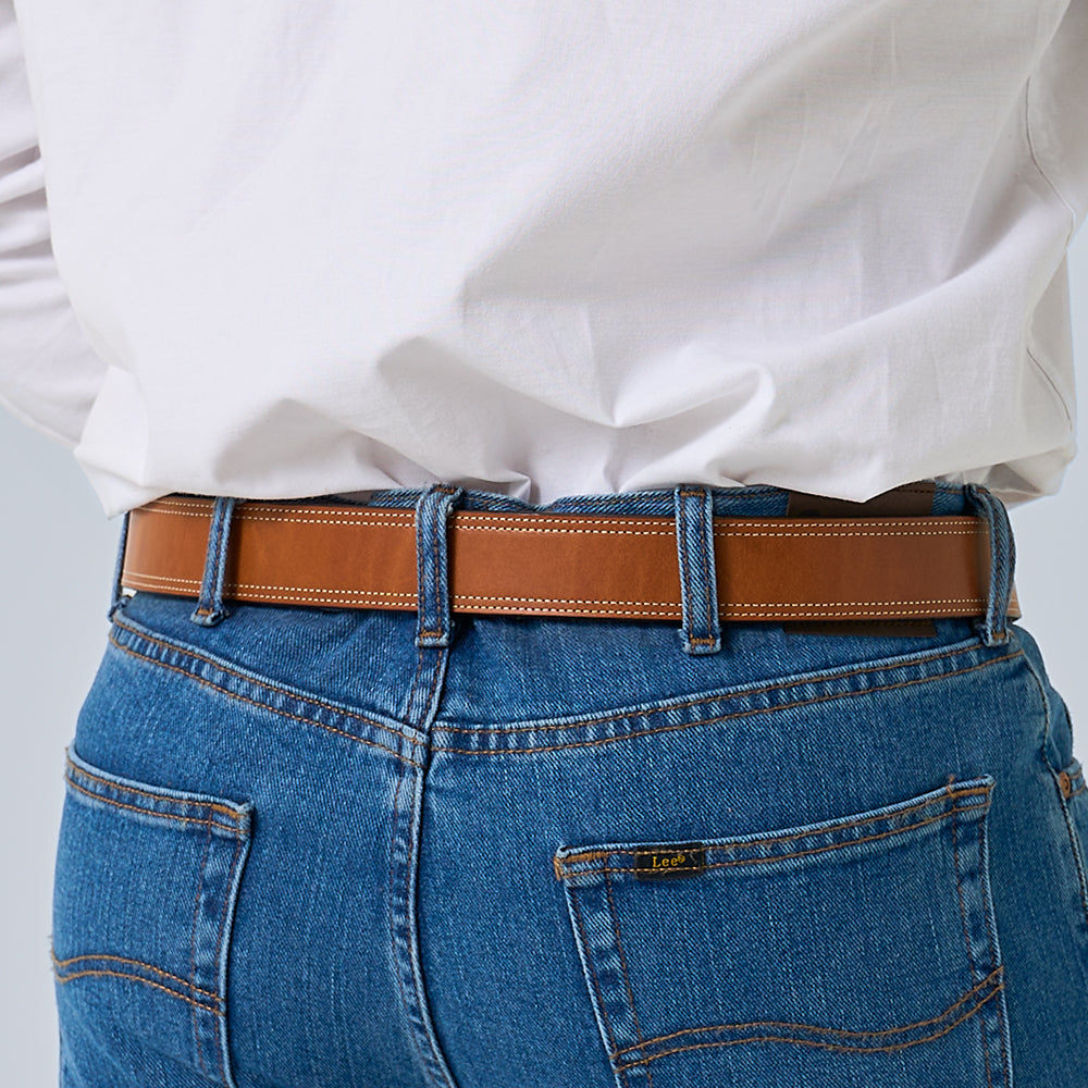 A person is shown from the back wearing blue denim jeans, a white shirt tucked in, and &quot;The Premier Double Layer&quot; leather belt by Hanks Belts; the jeans have visible stitching, belt loops, and a &quot;Lee&quot; brand label on the pocket.
