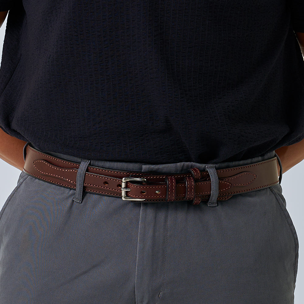 A person wearing gray pants and a dark navy shirt is shown with The Ranger belt by Hanks Belts—a brown full-grain leather accessory with decorative stitching and a silver buckle. The image highlights the waist where the belt is worn.