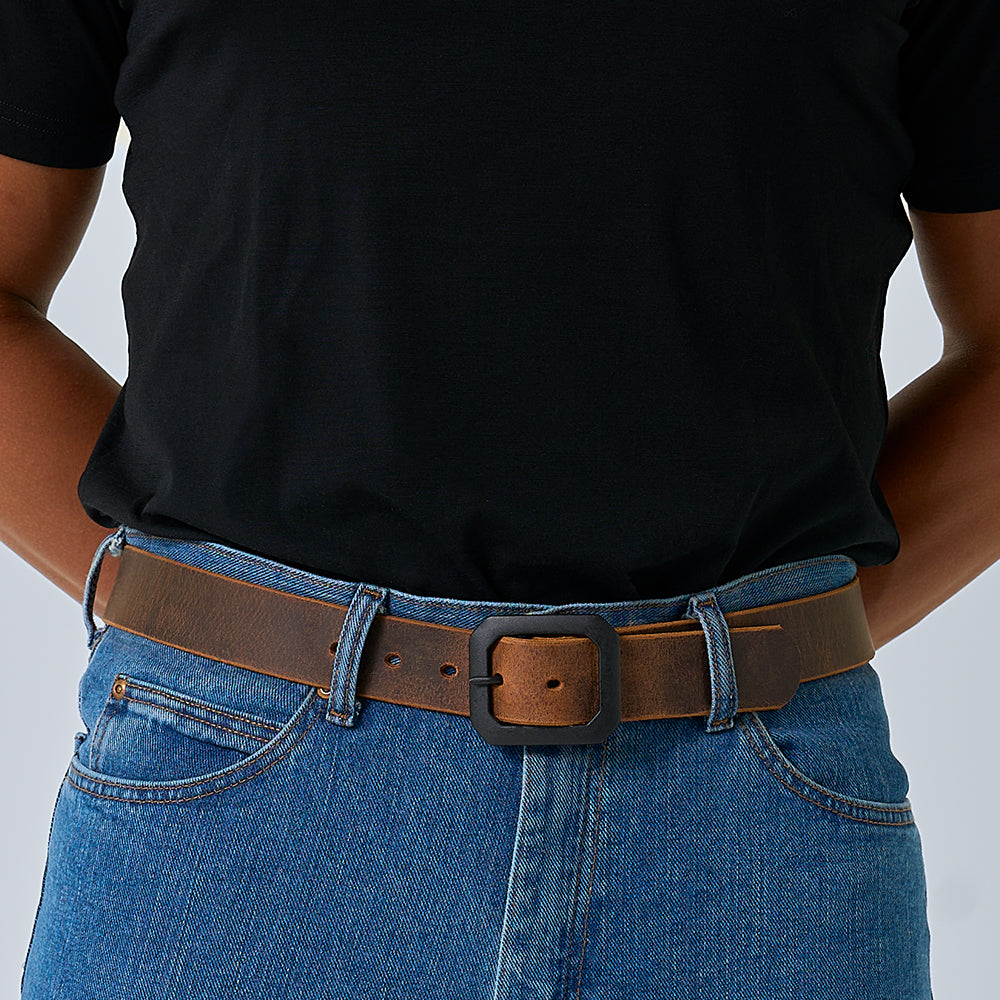 A person wears a black t-shirt tucked into blue jeans with The Relic Retro Style belt by Hanks Belts, featuring a large square buckle. Their hands are behind their back.