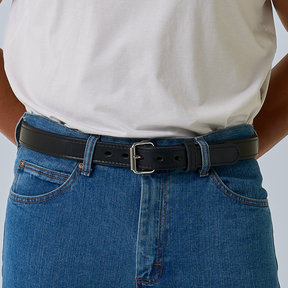 A person wearing a white T-shirt tucked into blue jeans is shown from the waist down, focusing on The Steel Core - 1.25&quot; black leather belt by Hanks Belts fastened around their waist.