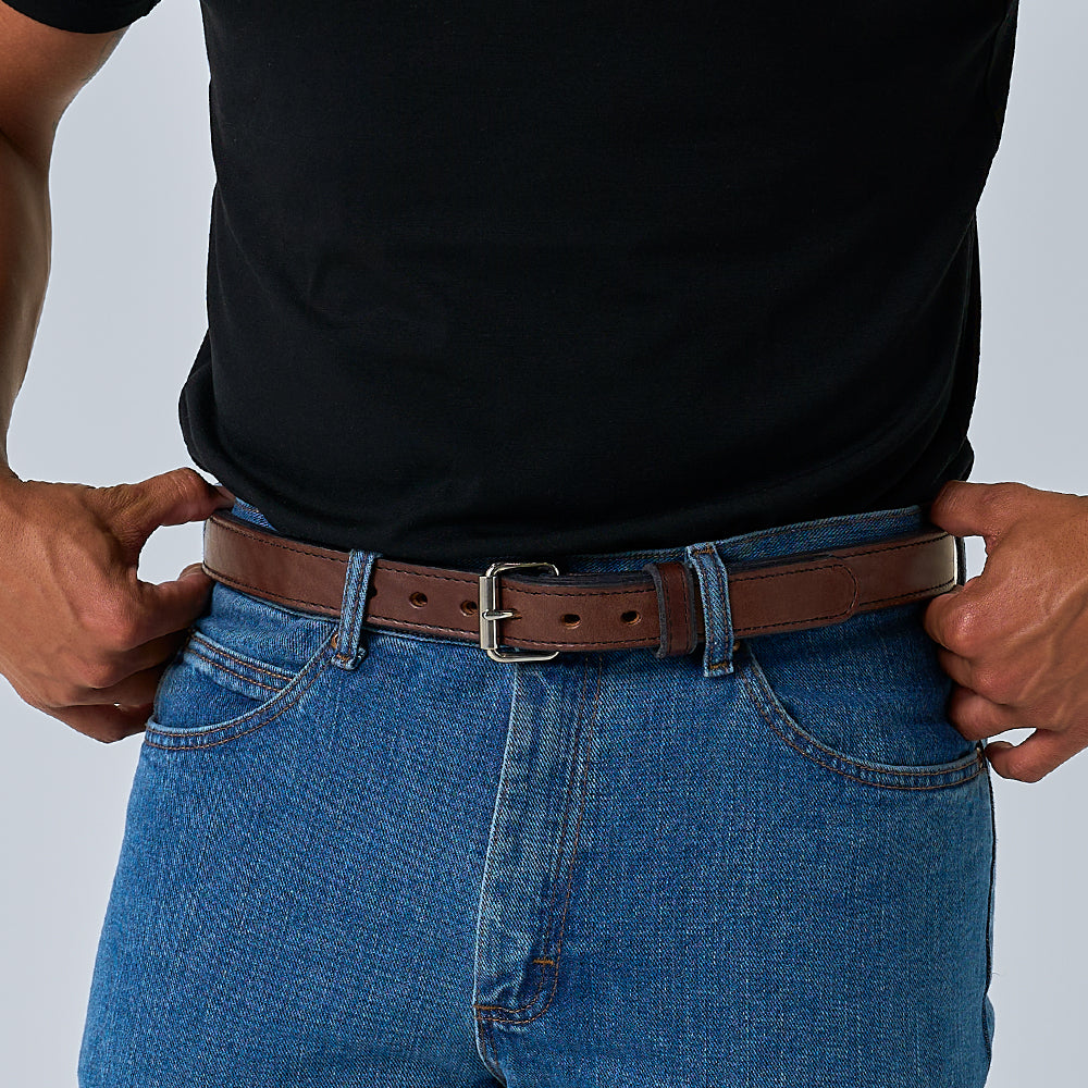 A person wearing a black t-shirt and blue jeans adjusts a Hanks Belts The Steel Core - 1.25&quot; brown leather belt around their waist, with only the torso and hands visible.