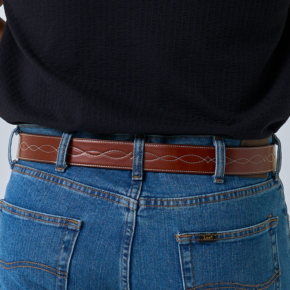 A person wearing blue jeans and a black top is shown from the lower back, featuring The Teton Tapered belt by Hanks Belts—a handcrafted leather accessory with decorative white stitching.